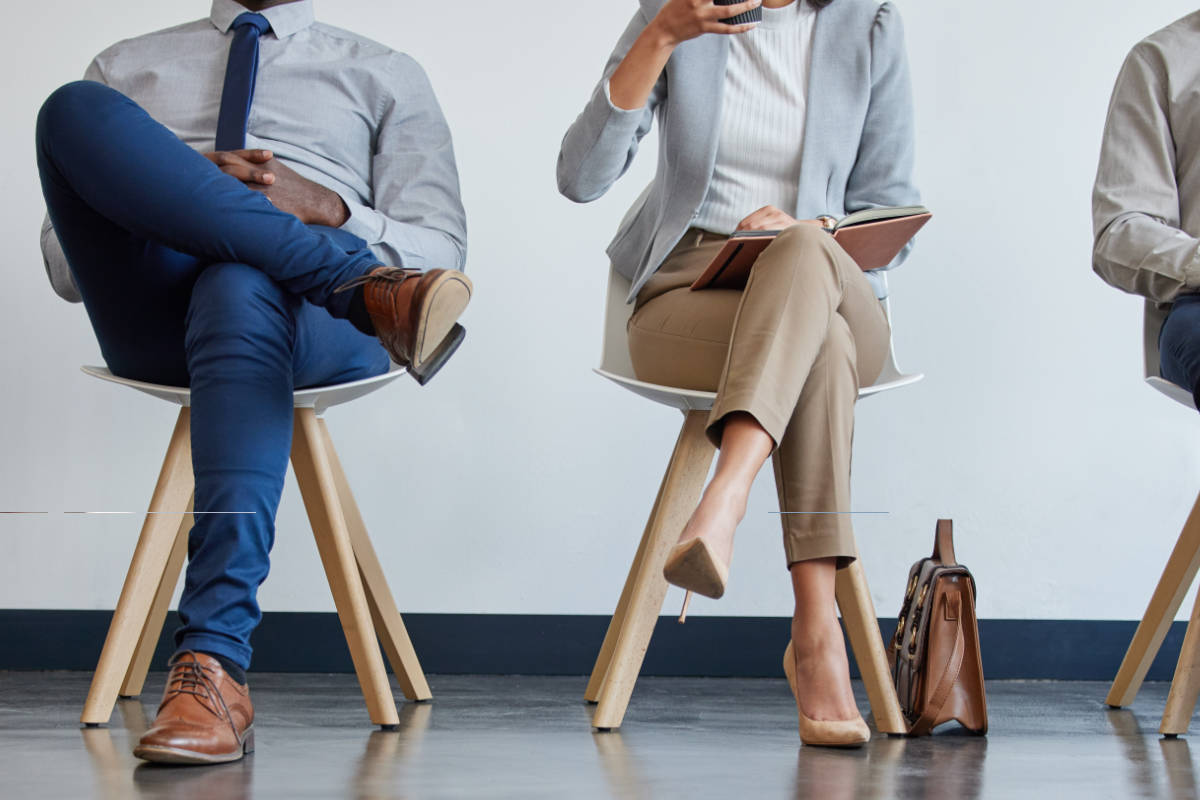 Vier Personen sitzen nebeneinander auf Stühlen in einem Wartebereich, teils mit Laptop, Notizbuch oder Kaffeebecher.