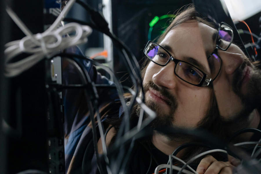 Person arbeitet hinter IT-Hardware zwischen Kabeln und Anschlüssen, konzentriert bei der technischen Wartung.